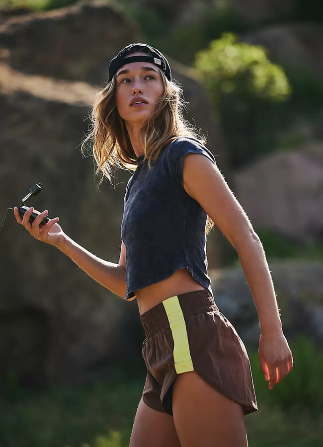 A young woman outdoors wears a black cap, a dark cropped T-shirt, and brown shorts with yellow stripes, effortlessly showcasing denim alternatives in current fashion trends. She holds a mobile or device, looking off to the side, with rocks and greenery in the background.