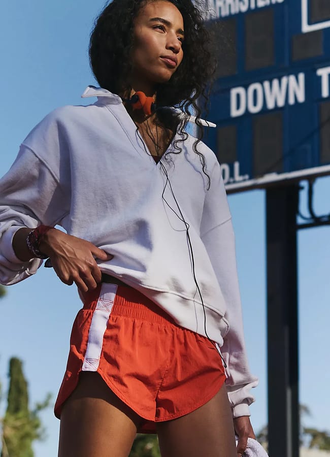 A person in a white jumper and red alternate shorts stands outdoors by a scoreboard, listening to music with wired earphones, with trees and blue sky in the background—a fresh take on summer fashion.