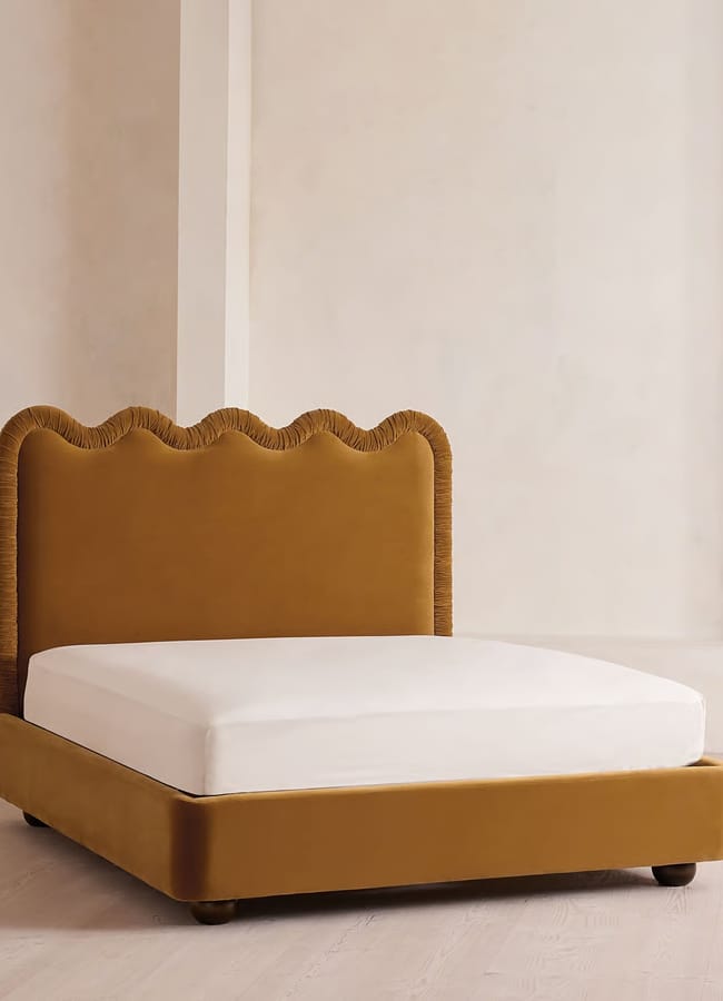 A modern bed with a mustard velvet frame and a wavy, scalloped headboard stands on a light wooden floor in a minimalist beige room, echoing the bold design sensibilities often found in maximalist hotels.