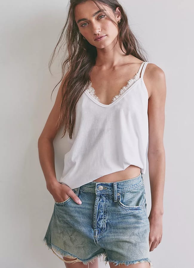 A woman with long brown hair wears a loose white vest top with lace trim and the best denim shorts—high-waisted and frayed—standing against a plain light-coloured wall with one hand in her pocket.