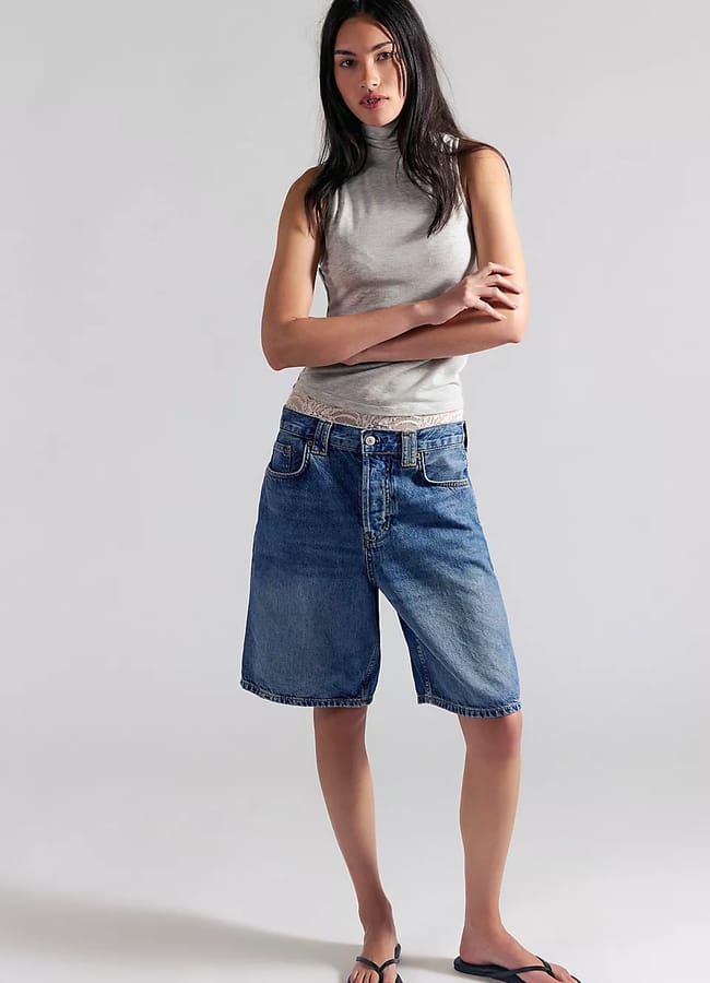 A woman stands against a plain background, wearing a sleeveless grey polo-neck top, best denim shorts in a loose knee-length fit, and black flip-flops. Her arms are crossed and her expression is neutral.