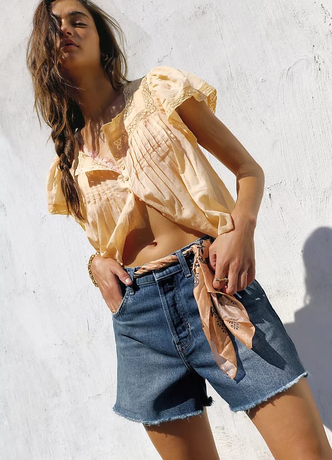 A woman poses outdoors against a white wall, wearing a light yellow blouse and the best denim shorts with frayed hems, accessorised with a peach scarf as a belt. She has long, wavy hair and wears rings on her fingers.