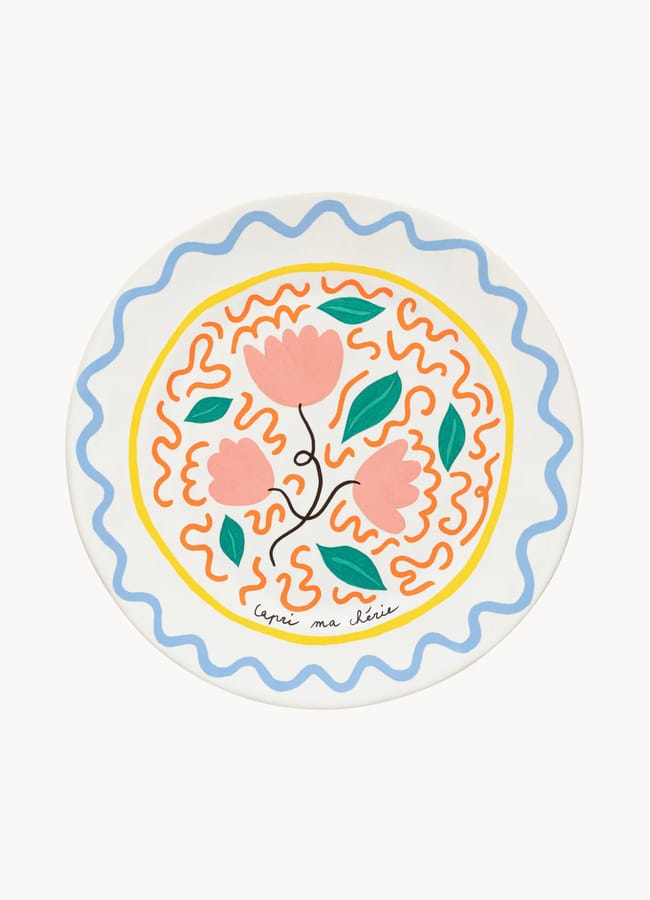 A round ceramic plate with a colourful, hand-painted design featuring pink flowers, green leaves, orange squiggles, and a wavy blue border—perfect for salad season. The artist’s signature is visible at the bottom.