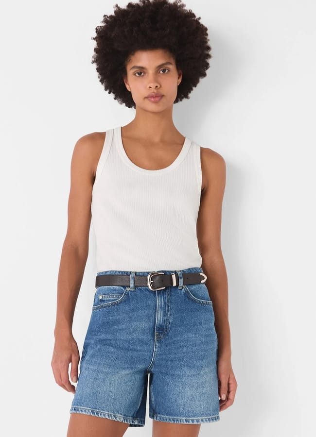 A person with short curly hair wears a white sleeveless ribbed vest, the best denim shorts, and a black belt, standing against a plain white background.