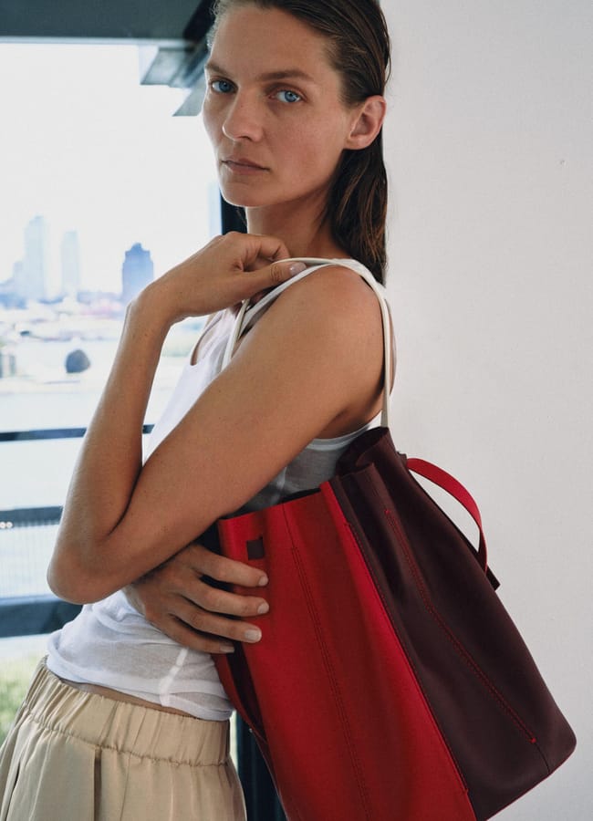A person with wet hair in a white vest and beige trousers stands indoors, holding a large red tote bag over their shoulder—a look that channels high street fashion. City buildings are visible through the window in the background.