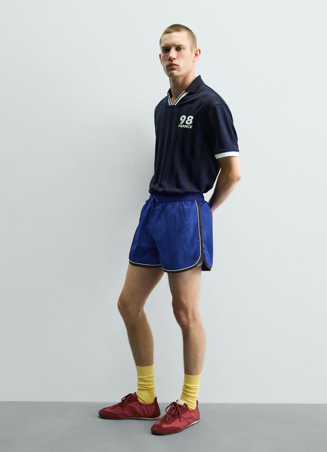 A person stands against a plain light-coloured wall, embodying football core style in a dark polo shirt with 98 FRANCE on it, blue athletic shorts, yellow socks, and red trainers. They have short hair and a neutral expression.