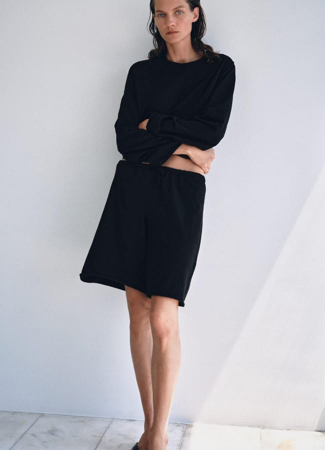 A person stands against a plain wall, embodying high street fashion in a loose black long-sleeve top, matching black shorts, and black slip-on sandals, with arms folded and a neutral expression.