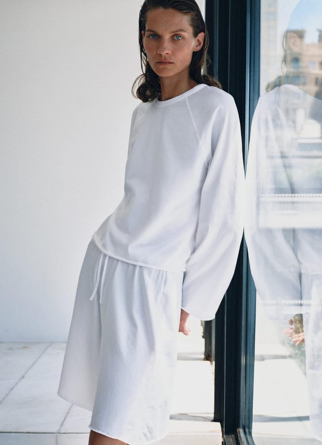 Person with wet, slicked-back hair wears a loose white long-sleeve top and white drawstring shorts, channelling high street fashion while standing indoors near a glass door with their reflection visible. The setting is minimal and bright.