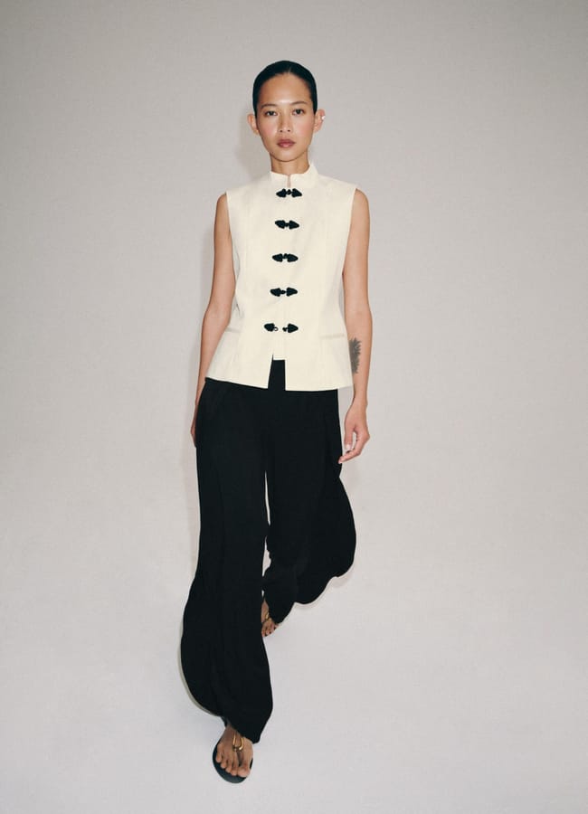 A person models a sleeveless, high-neck white top with black frog fastenings and loose black trousers against a plain background, highlighting the chic outfit’s striking fashion detail.