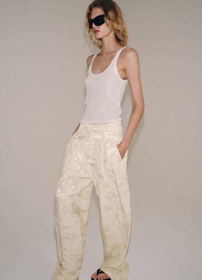 A person wearing a white vest, loose cream-coloured floral trousers, black sandals, and dark sunglasses stands against a plain light backdrop with hands in pockets, effortlessly embodying high street fashion.
