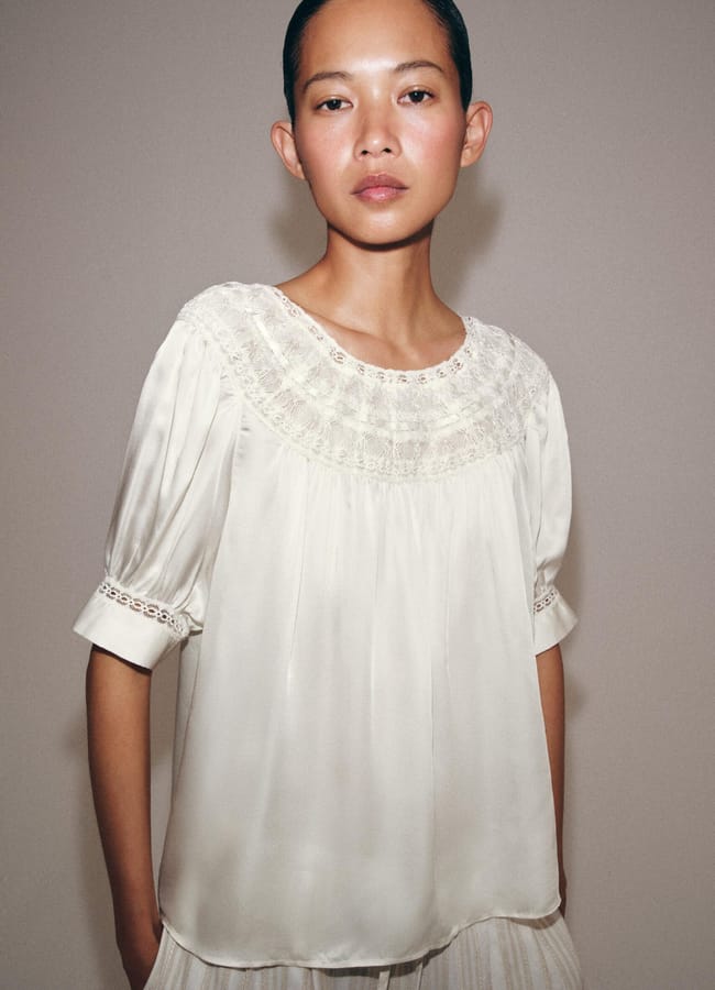 A person with short dark hair wears a cream-coloured, short-sleeved, loose blouse with lace details and pleats—a refined take on high street fashion—standing against a plain light background.