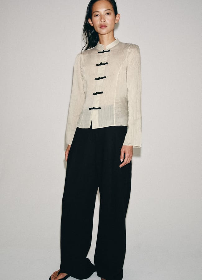 A person stands against a plain background wearing a beige, long-sleeved, mandarin-collar jacket with black frog fastenings—a subtle fashion detail—paired with loose black trousers and black sandals.