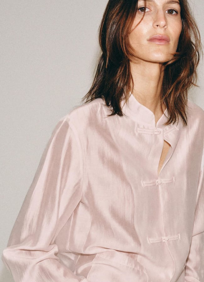 A person with medium-length brown hair wears a light pink, silk blouse with a mandarin collar and decorative frog fastenings, showcasing exquisite fashion detail while standing against a plain background.