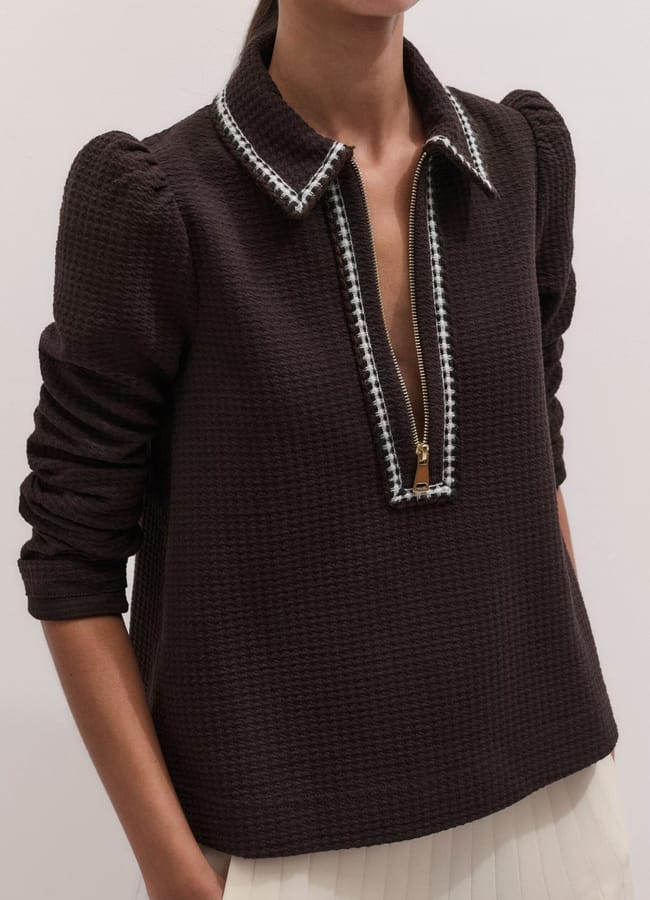 A person wearing a textured dark brown top with a deep zippered neckline, puffed sleeves, and a white-trimmed collar, paired with a pleated white skirt—a chic ensemble that perfectly captures high street fashion. Their face is not visible.