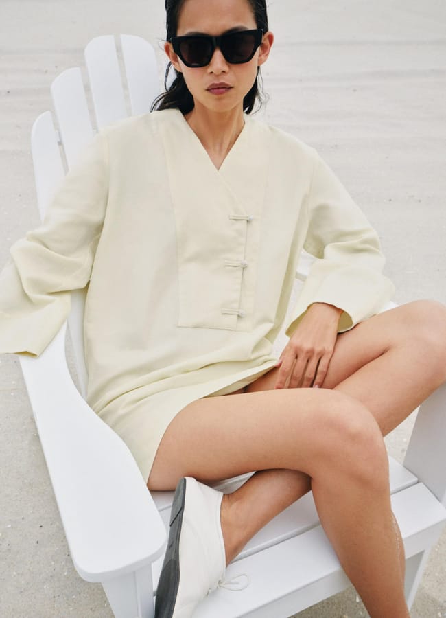 Woman in a light cream tunic featuring subtle fashion detail and sunglasses sits on a white Adirondack chair on a sandy beach, legs crossed, with relaxed posture and neutral expression.
