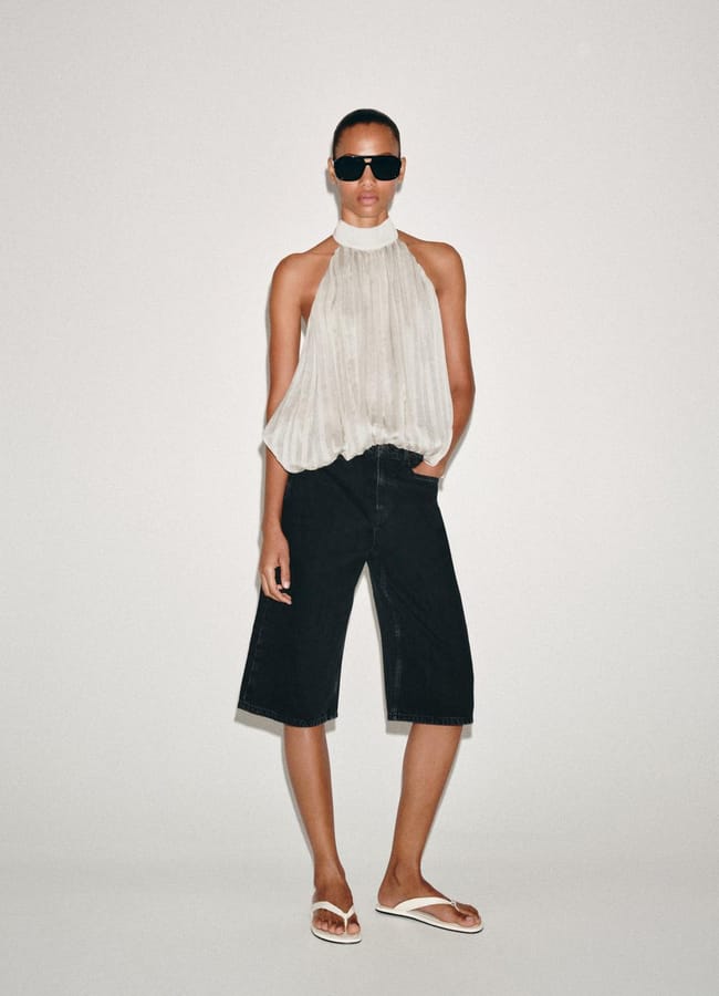 A person wearing a sleeveless, high-neck white pleated top, best denim shorts, white flip-flops, and black sunglasses stands against a plain light background with one hand in their pocket.