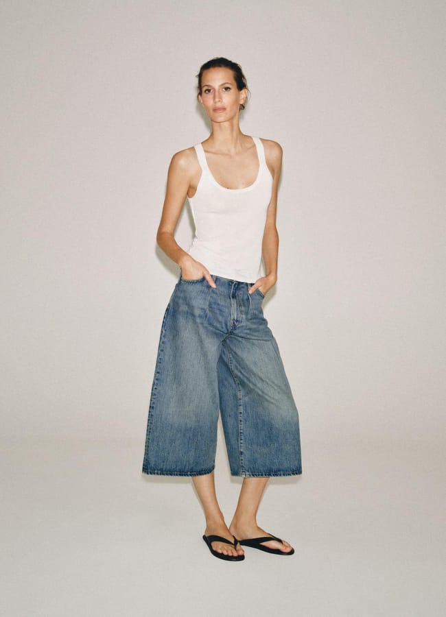 A person stands against a plain background wearing a white vest, wide-leg blue denim culottes styled like the best denim shorts, and black flip-flops, with hands in their pockets and a relaxed expression.