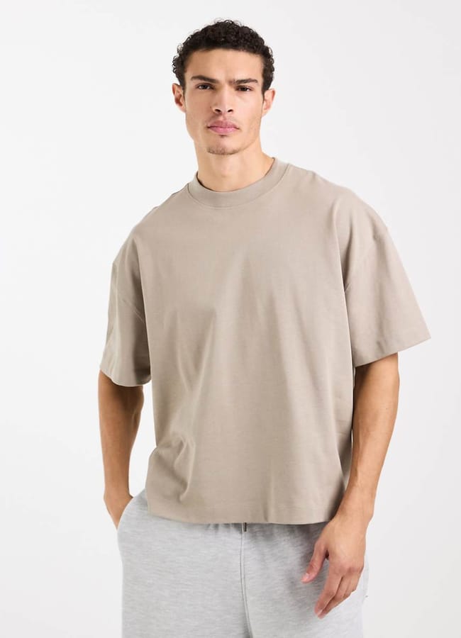 A person wearing a loose beige t-shirt and light grey tracksuit bottoms stands against a plain white background, channelling understated celebrity fashion while looking at the camera with a neutral expression.