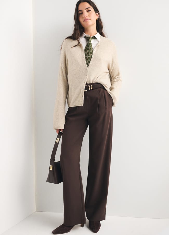 Woman stands against a white wall wearing a beige cardigan, white shirt, patterned green tie, brown high-waisted trousers, brown heels, and holding a matching brown handbag—a refined look that captures the essence of autumn winter 2026 fashion trends.