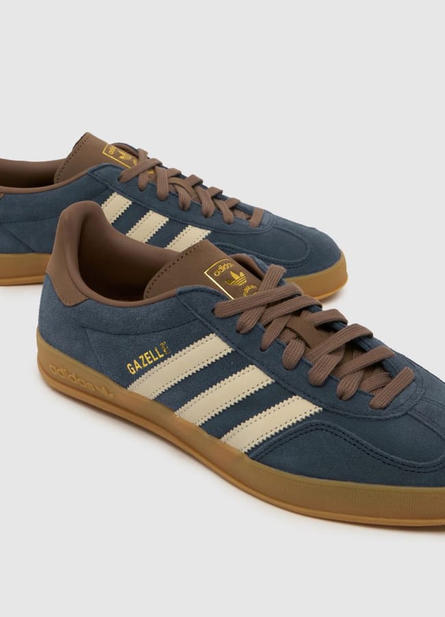 A pair of blue Adidas Gazelle trainers with white stripes, brown laces, and gum soles are displayed on a plain light grey background—an effortless touch of celebrity fashion perfect for elevating your holiday style.