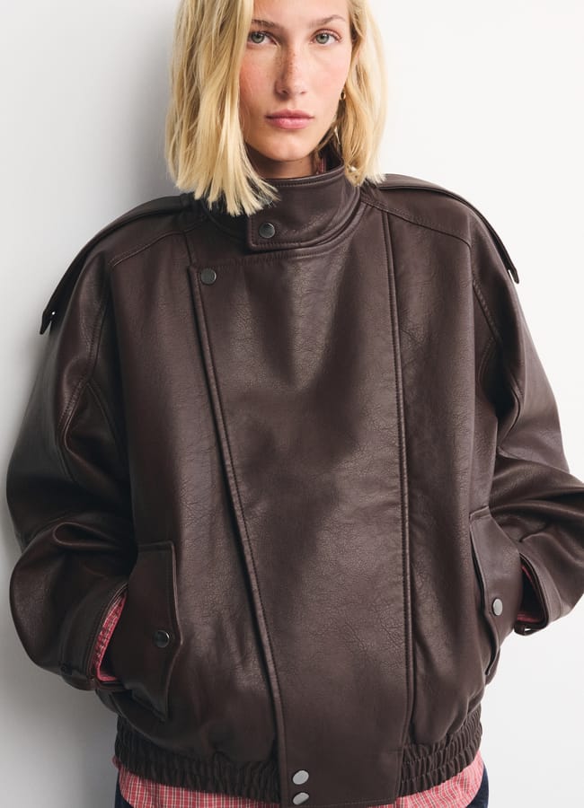 A woman with blonde hair wears a loose-fitting dark brown leather jacket over a pink checked shirt, standing against a plain white wall with her hands in her pockets—a stylish look that captures the essence of autumn winter 2026 fashion trends.