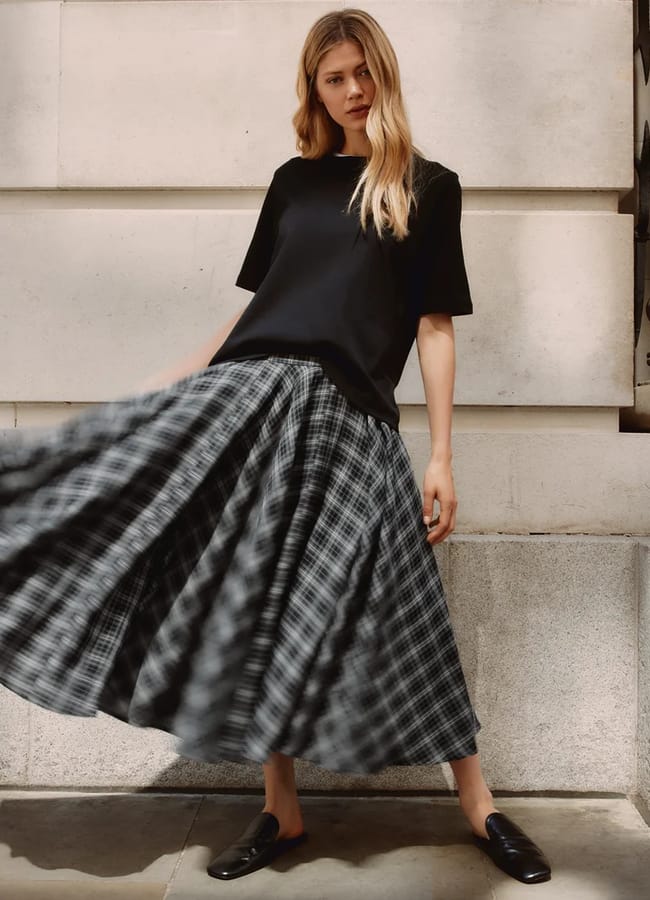 Woman wearing a black T-shirt and a flowing black-and-white checked skirt stands against a stone wall, with one foot forward and skirt swaying, creating a dynamic movement. Embracing autumn winter 2026 fashion trends, she has long blonde hair and wears sleek black shoes.