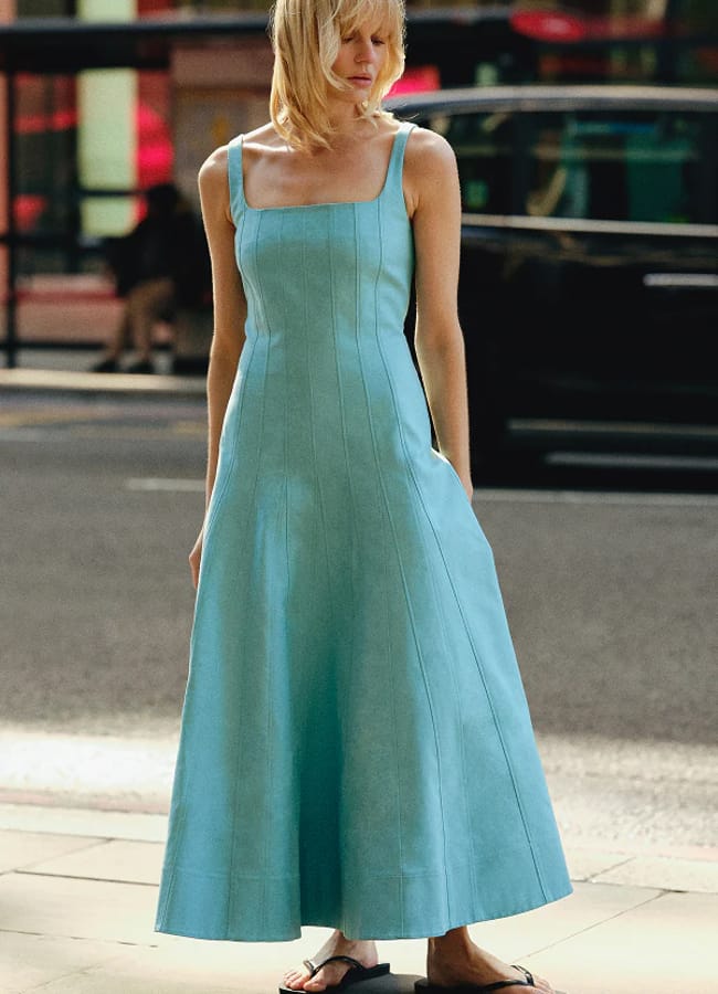 A person with blonde hair wears a sleeveless, light blue, square-neck, ankle-length dress—showcasing a touch of turquoise fashion—and black sandals, standing on a city pavement with a street and vehicles in the background.