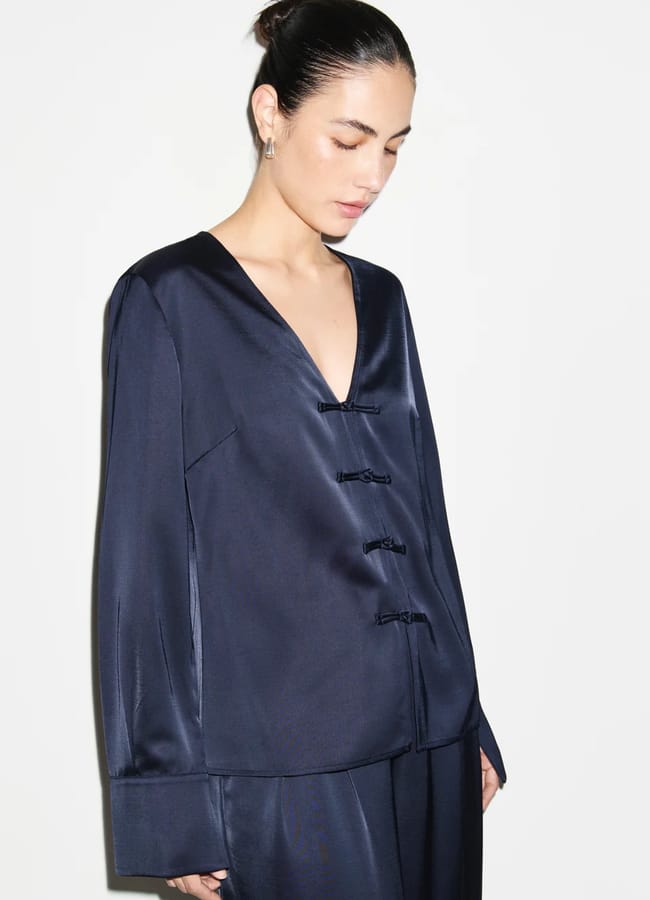 A woman with dark hair in a bun wears a navy satin blouse with a deep V-neck, long sleeves, and three decorative knots on the front—an elegant piece perfectly in line with autumn winter 2026 fashion trends—standing against a plain white background.