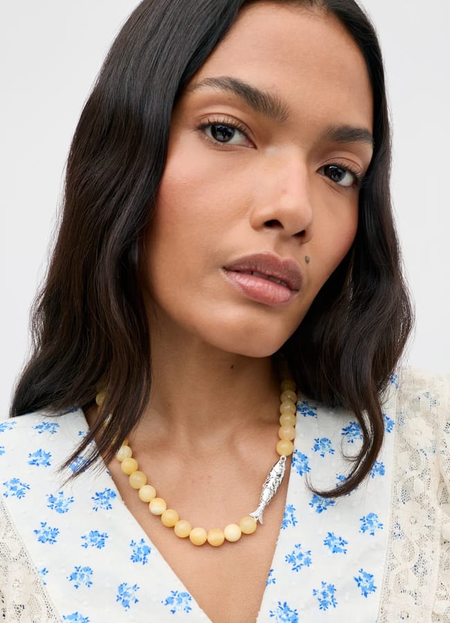 A woman with long dark hair wears a cream beaded necklace with a silver feather pendant, showcasing her taste in fashion accessories. She has a neutral expression and is dressed in a white top with blue floral patterns and lace trim.