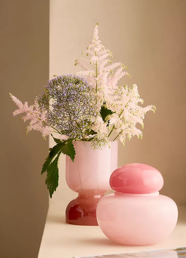 A pink glass vase, one of the statement vases in the scene, with delicate purple and pale pink flowers sits beside a round pink glass jar on a beige surface, with a magazine partially visible in the foreground.
