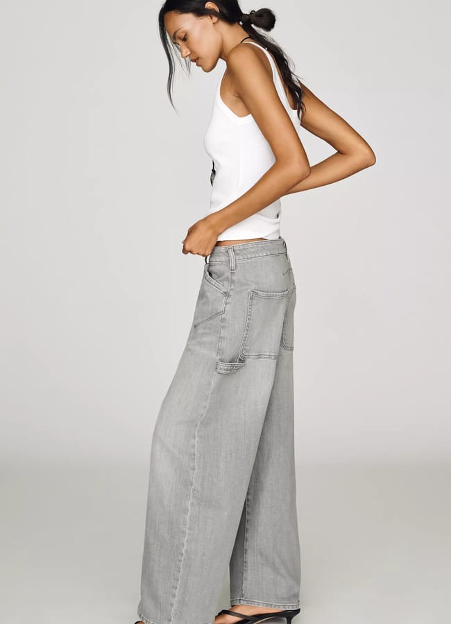 Woman in a white vest top, grey wide-leg jeans, and black heeled sandals stands sideways against a plain background, looking down with one arm slightly bent and her hair tied back—a subtle nod to effortless fashion. For the coolest way to elevate this look, consider adding an animal print accessory for a bold yet chic touch.
