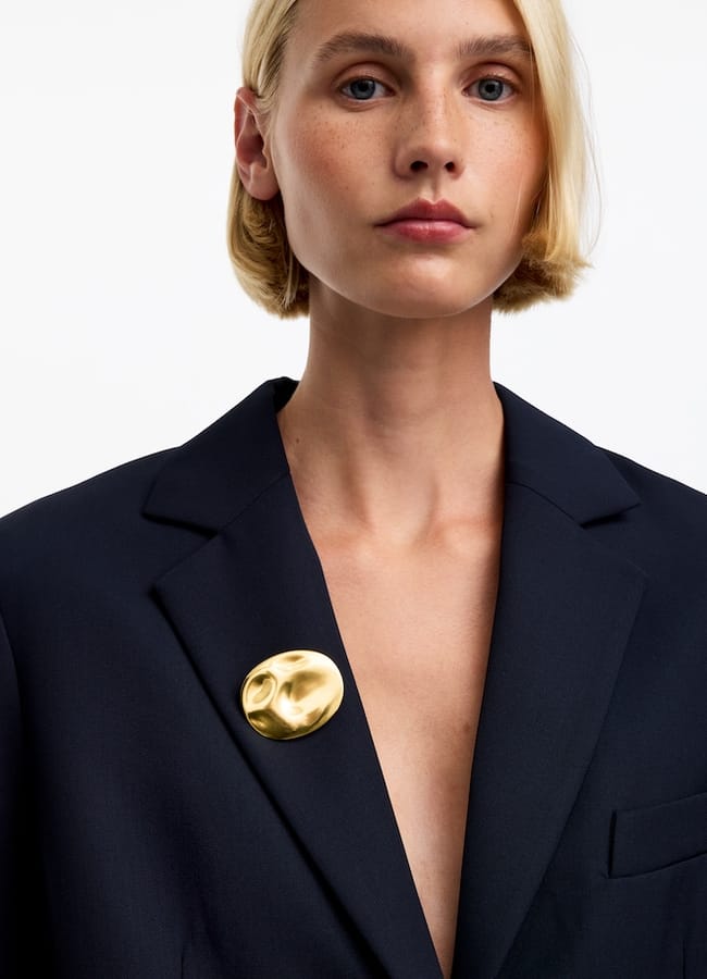 A person with short blond hair, embodying editor fashion, wears a dark blazer with a large, round, gold pin on the lapel. The background is plain white.