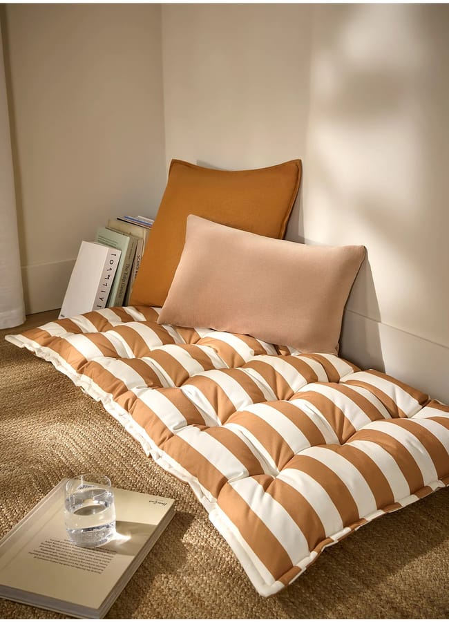 A cosy reading nook with a striped floor cushion, two pillows, a stack of books, an open book, and a glass of water on a woven rug by the corner of a sunlit room—perfect for unwinding after an at-home film night.