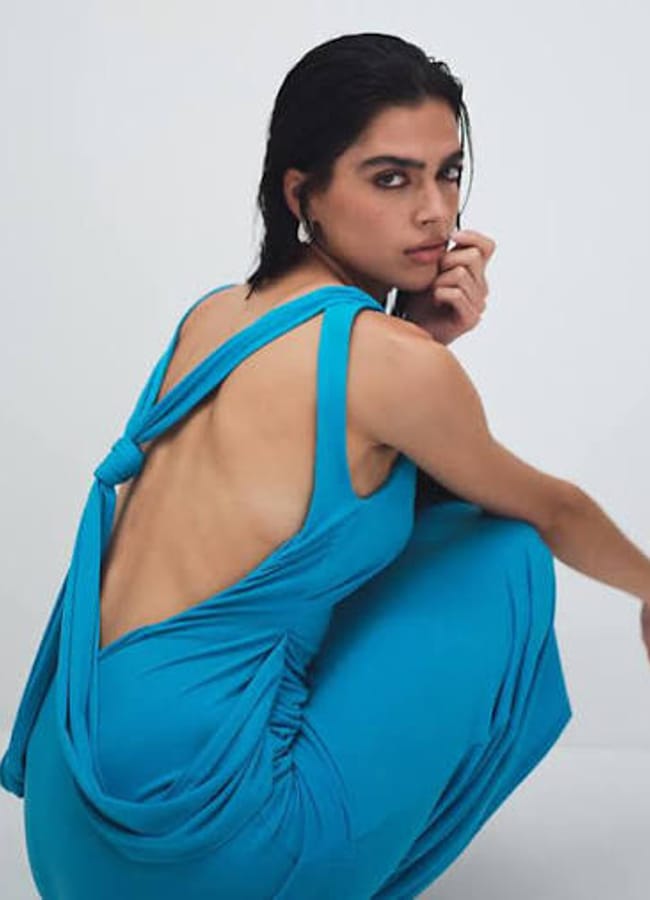 A woman with wet, dark hair squats in a bright blue, backless dress with draped straps, looking over her shoulder against a plain, light background—a striking nod to street style inspired by the latest fashion trends.