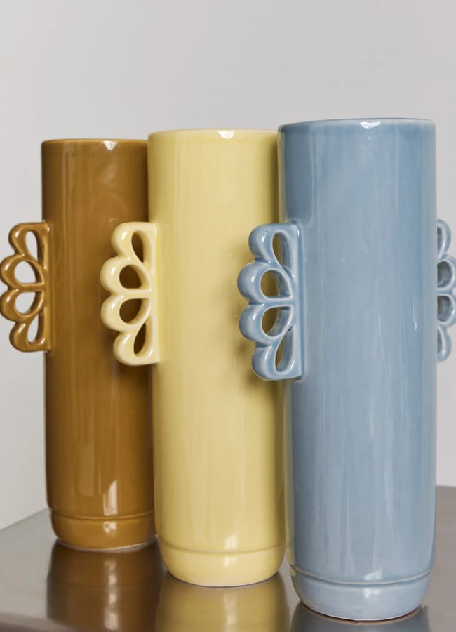 Three tall ceramic statement vases in brown, yellow, and blue stand side by side on a wooden surface. Each vase features a decorative handle with a petal-like cut-out design.