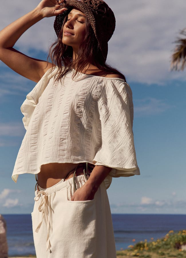 A woman in a white, loose outfit and brown sunhat stands outdoors near the sea, eyes closed, enjoying the tranquil breeze of her last-minute holiday, with one hand holding her hat under a partly cloudy sky.