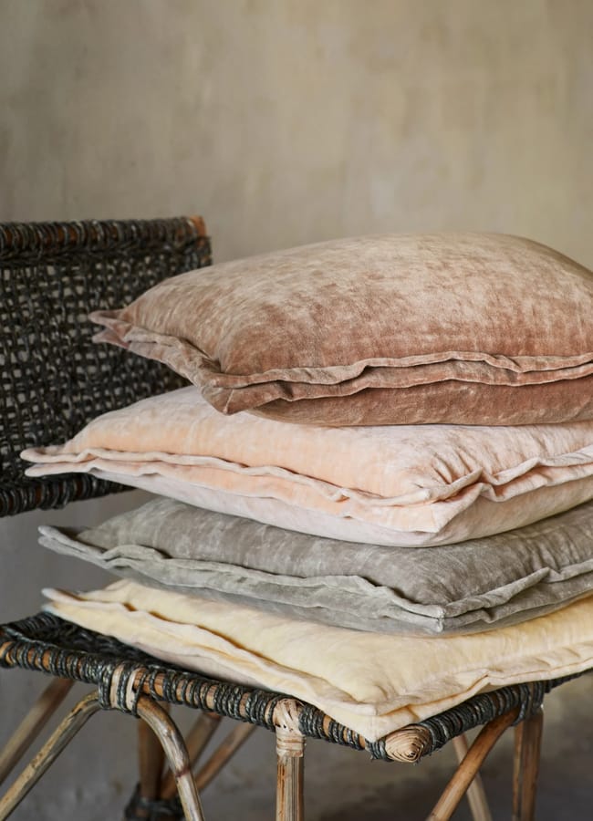 Four soft, pastel-coloured cushions in shades of brown, pink, grey, and yellow are stacked on a woven chair against a neutral, textured wall background—perfect for creating a cosy atmosphere during your next outdoor-film-night-at-home.