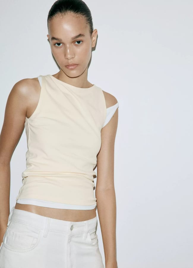 A person with slicked-back hair wears a sleeveless beige top over a white vest and white trousers, standing against a plain white background—one of our favourite fashion picks for effortless holiday style.