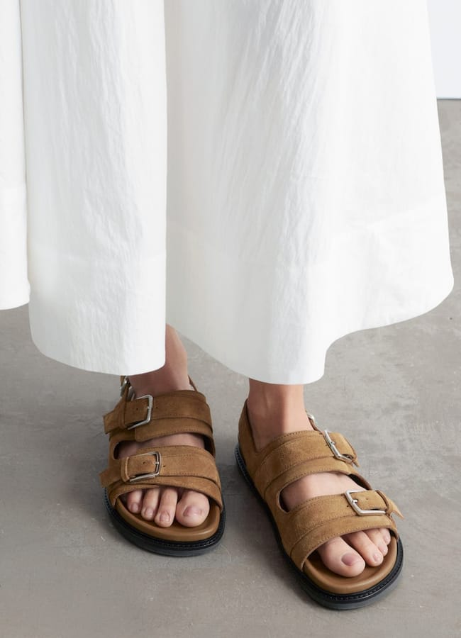 Person wearing a long white skirt and tan suede sandals with double buckles, standing on a concrete floor—perfect inspiration for effortless holiday fashion. Only the lower legs and feet are visible.
