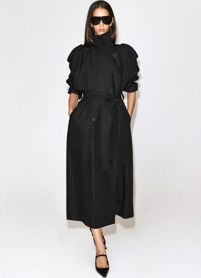 A woman wearing a long black trench coat, black heels, and large black sunglasses stands against a plain white background with hands in her coat pockets, embodying the sleek sophistication of autumn winter 2026 fashion trends.