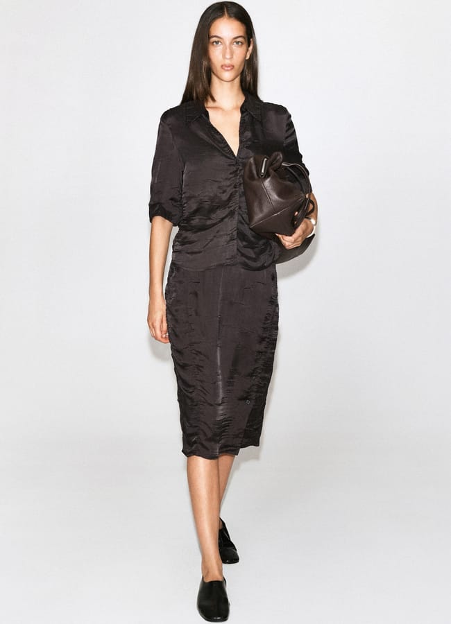 Woman wearing a shiny black blouse and skirt, holding a large brown leather bag, walking forward against a plain white background, with black shoes and straight brown hair—a look that perfectly captures the essence of autumn winter 2026 fashion trends.