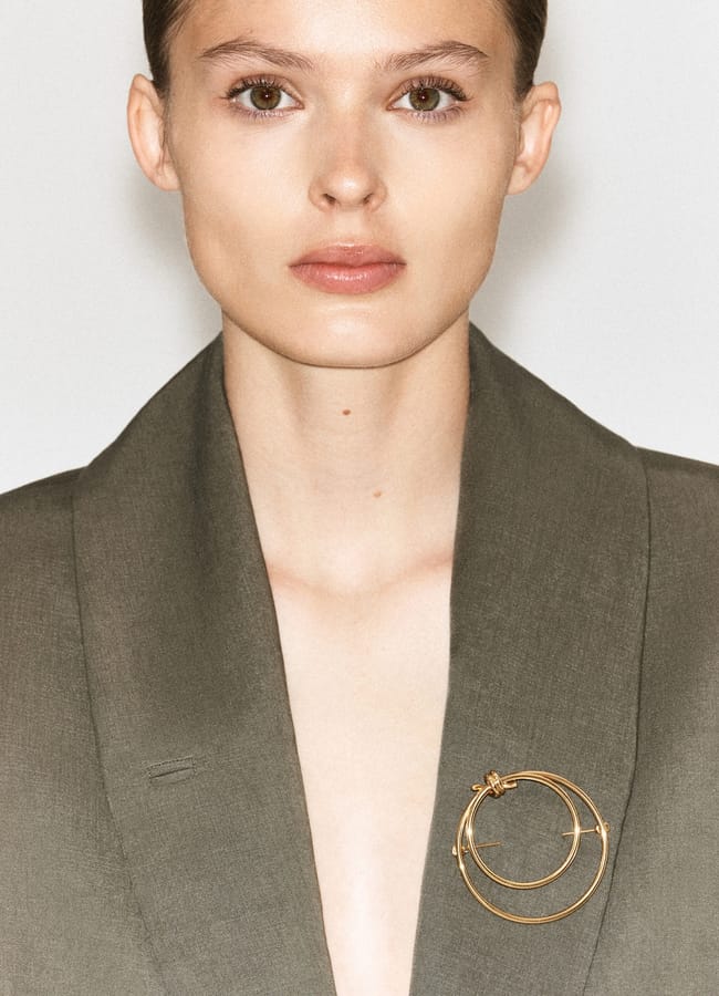 A person wearing an olive green blazer with a large gold circular brooch on the lapel, pictured from the chest up against a plain background—an elegant look that perfectly reflects autumn winter 2026 fashion trends.