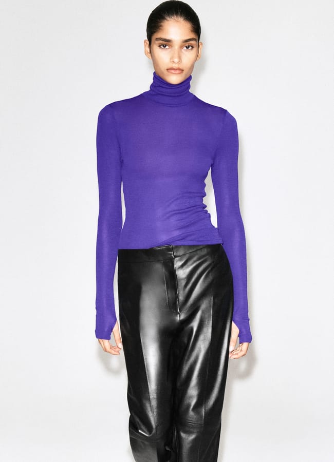 A person wearing a fitted, long-sleeve purple polo neck and black leather trousers stands against a plain white background, looking directly at the camera—a striking look that perfectly captures autumn winter 2026 fashion trends.