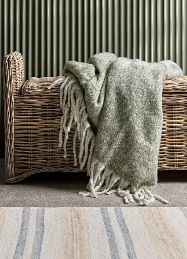 A green and white fringed throw blanket draped over a woven wicker bench, perfect for cosying up during an outdoor-film-night-at-home, with a ribbed green wall and a striped rug in the background.