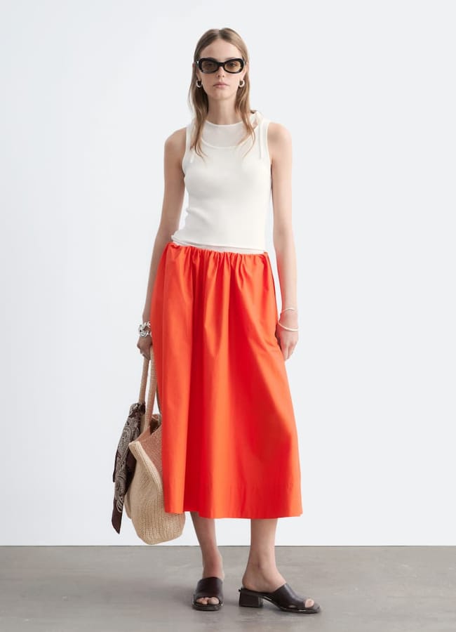 A woman, embodying holiday fashion, wears sunglasses, a sleeveless white top, and a bright orange midi skirt as she stands holding a straw bag. She completes her look with black sandals and a brown scarf. The background is plain and light-coloured—perfect inspiration for your last minute holiday fashion pics.