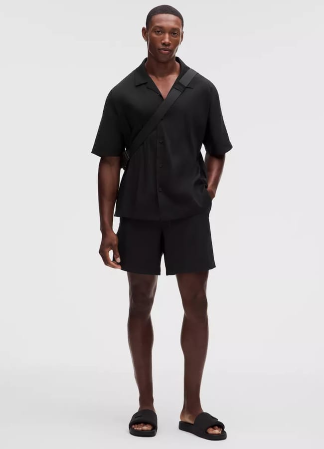Man stands against a plain background wearing a black short-sleeved shirt, black shorts, black slide sandals, and a black crossbody bag—exuding effortless style reminiscent of celebrity couples on holiday. One hand is in his pocket and he is looking at the camera.