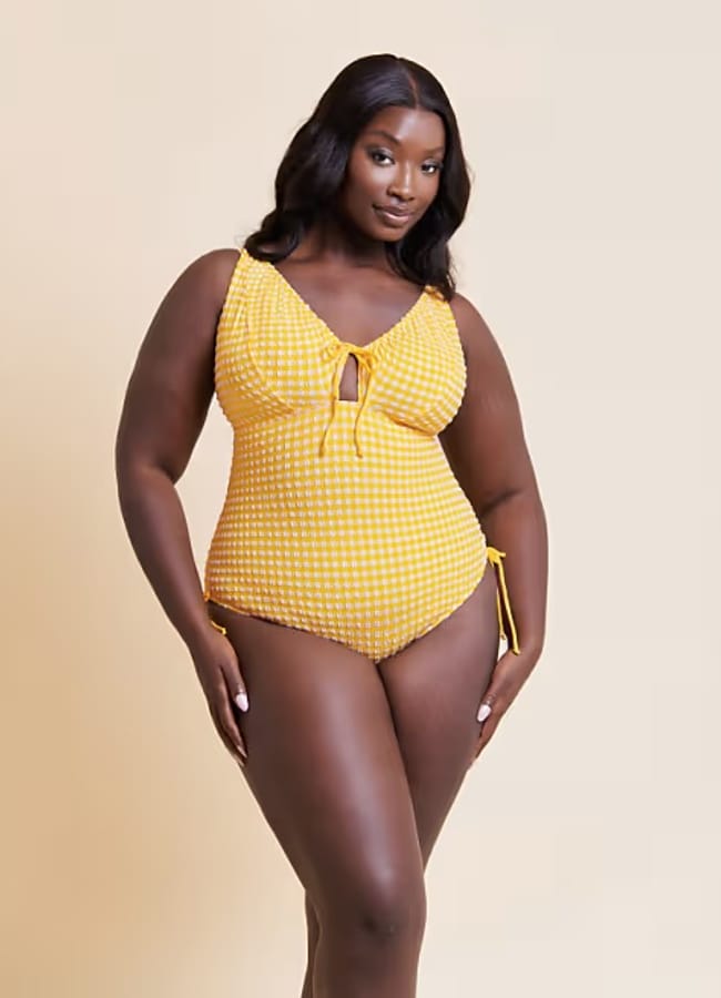 A woman wearing a pretty yellow and white chequered one-piece swimming costume poses confidently against a plain beige background, showing off her stylish swimwear.