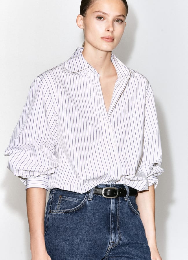 A person wearing an oversized white, long-sleeved, collared shirt with thin vertical stripes and blue jeans stands against a light background with a neutral expression, offering subtle inspiration for oversized shirt outfit ideas.