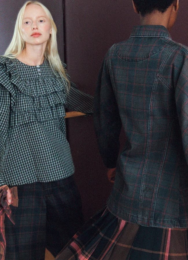 Two women showcase autumn winter 2026 fashion trends in checked clothing as they stand indoors; one faces forward in a frilled top, holding a bag, while the other turns away in a coordinating checked shirt and skirt. Both have light skin, and the maroon background adds warmth to this stylish seasonal scene.