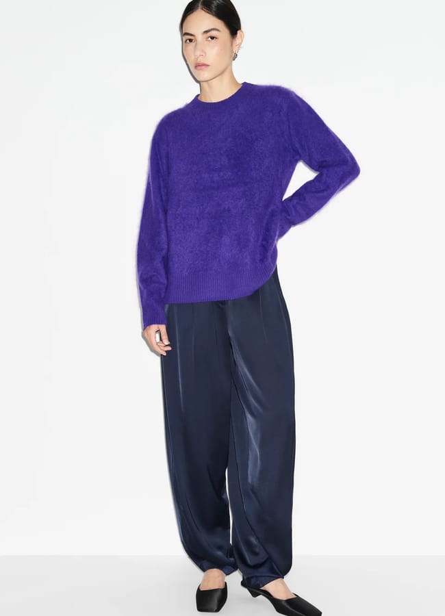 Person standing against a white background, wearing a fluffy purple jumper, loose dark navy trousers, and black slip-on shoes, with one hand resting on their hip—a look embodying autumn winter 2026 fashion trends.